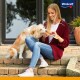 Une femme en cardigan bordeaux est assise sur des marches en pierre, tenant un paquet de friandises pour animaux de compagnie jaunes, tandis qu'un golden retriever s'appuie contre elle.