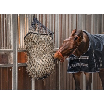 Ein braunes Pferd, das mit einer schwarzen Decke gefüttert wird, frisst von einem Heunetz, das in seiner Box hängt, umgeben von Holzwänden und Metallstangen.