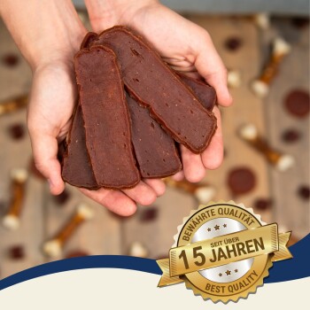 Hände halten mehrere Streifen von Hundeleckerlis, mit einem Abzeichen, das "15 Jahre" und "Beste Qualität" in der Ecke angibt.