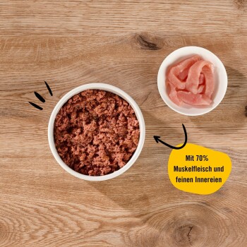 Haustierfutter in einer weißen Schüssel, mit gehacktem Fleisch und einer Seite von rosa Fleischscheiben, beschriftet 'Mit 70% Muskelfleisch und feinen Innereien.'