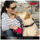 A woman in a striped shirt sits outdoors with a yellow Labrador, holding a red Flexi retractable leash. The background features wooden structures.