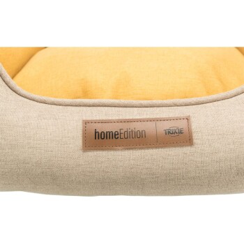 Eine Nahaufnahme eines Haustierbettes mit einem beigen Stoffäußeren und einem gelben Kissen, mit einem Lederetikett, das 'homeEdition' und 'TRIXIE' liest.