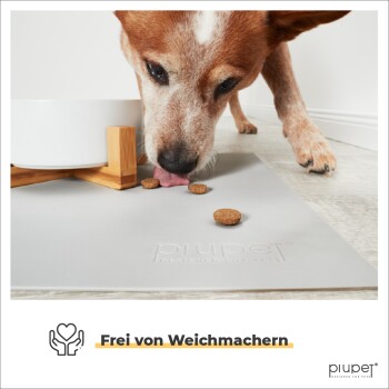 Eine Nahaufnahme eines Hundes, der Trockenfutter von einer grauen Matte frisst, mit einer weißen Schüssel auf einem Holzständer in der Nähe. Der Text lautet "Frei von Weichmachern."