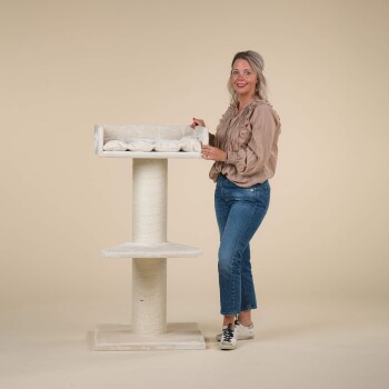 Eine Frau steht neben einem hohen, weißen Katzenbaum mit einer plüschigen oberen Plattform und einem Kratzbaum, trägt eine beige Bluse und blaue Jeans.