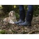 Eine Person in stilvollen schwarzen und gemusterten Gummistiefeln steht auf einem Waldboden und schaut auf einen flauschigen Hund neben sich.