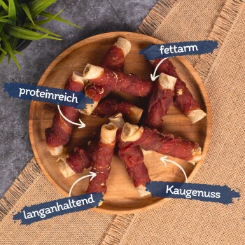 Ein Holzbrett zeigt gerollte Hundeleckerlis, die mit "hoher Proteinanteil", "wenig Fett", "langanhaltend" und "Kaugenuss" beschriftet sind.