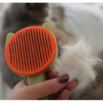 Eine Hand, die eine Katzenpflegebürste mit einem orangefarbenen Kopf und einem grünen Griff hält, neben einem Klumpen weißem Fell.