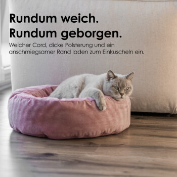 Graue Katze liegt in einem runden, rosa Cord-Katzenbett auf Holzfußboden; Text im Bild: 'Rundum weich. Rundum geborgen.'