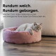 Graue Katze liegt in einem runden, rosa Cord-Katzenbett auf Holzfußboden; Text im Bild: 'Rundum weich. Rundum geborgen.'
