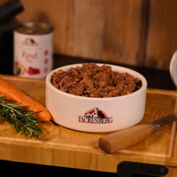 Weiße Keramikschüssel mit Tacrenberg-Logo gefüllt mit braunem Tierfutter, Karotte, Rosmarin und Messer auf Holzschneidebrett.