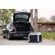 Ein schwarzes Auto mit offenem Kofferraum, geparkt neben einer tragbaren Haustierbox mit Netzseiten, vor einer Kulisse aus Bäumen und einer Holzstruktur.