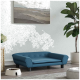 Ein stilvolles blaues Haustiersofa mit Knopfdetails, platziert neben einer weißen Futterschüssel, die mit Trockenfutter gefüllt ist, vor einer hellen Wand und einer Pflanze.