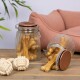 Zwei Glasbehälter gefüllt mit Hundeknochen, einer umgekippt, neben zwei Seilspielzeugen auf einer Holzoberfläche, mit einem rosa Haustierbett im Hintergrund.