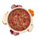 Weiße Schale mit stückigem Nassfutter für Haustiere mit sichtbaren Fleischstücken, umgeben von Birnen-, Apfel- und Karottenscheiben sowie rohem Fleisch.