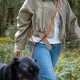 Person in beiger Jacke und Jeans geht mit einem schwarzen Hund an einer orangefarbenen freihändigen Leine in einem Wald mit gelben Blumen spazieren.