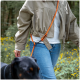Person in beiger Jacke und Jeans geht mit einem schwarzen Hund an einer orangefarbenen freihändigen Leine in einem Wald mit gelben Blumen spazieren.