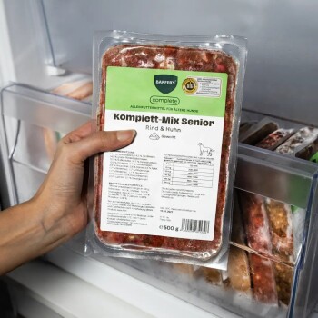 Hand hält vakuumversiegelte 500 g Hunde‑Futterpackung mit der Aufschrift 'BARFERS complete Komplett‑Mix Senior Rind & Huhn' im Gefrierschrank.