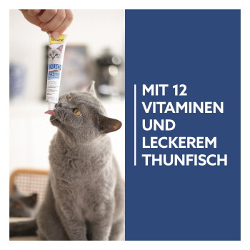Graue Katze leckt GimCat Duo Paste aus einer handgehaltenen Tube neben blauem Feld mit Aufschrift 'MIT 12 VITAMINEN UND LECKEREM THUNFISCH.'