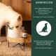 Ein weißer Hund frisst aus einer Schüssel auf einem Holzständer, neben einer Flasche mit Nahrungsergänzungsmitteln mit Dosierungs- und Lagerungshinweisen.
