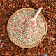 Eine Holzschüssel gefüllt mit bunten Haustierfutterpellets in Pink, Grün und Weiß, begleitet von einem kleinen Löffel, vor einem strukturierten braunen Hintergrund.