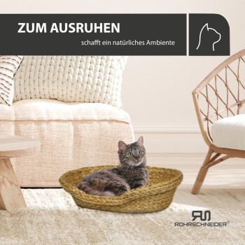 Eine getigerte Katze entspannt sich in einem geflochtenen Korbbett in einem gemütlichen Wohnzimmer mit einem hellen Sofa und Kissen. Text: "ZUM AUSRUHEN schafft ein natürliches Ambiente."