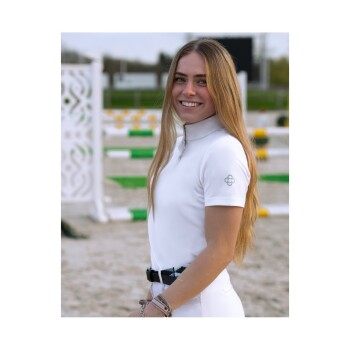 Eine Person in einem weißen Reitoutfit steht in einer Außenreitbahn mit grünen und gelben Sprüngen im Hintergrund.