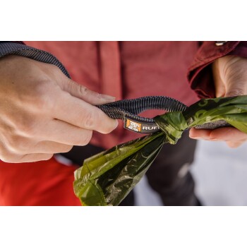 Eine Person hält eine Hundeleine mit einem 'Ruffwear'-Etikett, während sie auch einen gebundenen grünen Müllsack greift, vor einem verschwommenen, schneebedeckten Hintergrund.