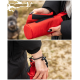 Eine Person hält einen roten Spender für Hundekotbeutel mit einem schwarzen Riemen, während ein kleiner Hund im Hintergrund unscharf ist.