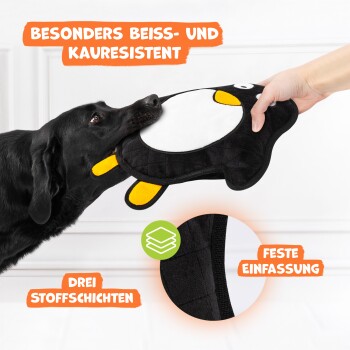 Ein schwarzer Hund, der verspielt an einem plüschigen Pinguinspielzeug zieht, mit dem Text 'Besonders Beiss- und Kauerresistent' und 'Drei Stoffschichten.'