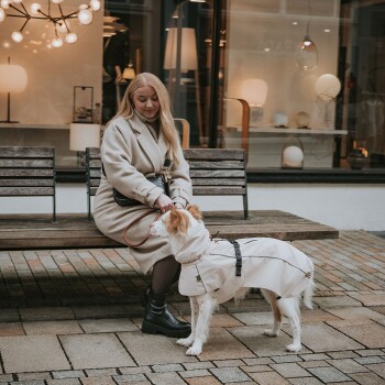 Eine Frau in einem beigen Mantel sitzt auf einer Holzbank und hält eine Leine, die an einem Hund mit einem weißen Regenmantel befestigt ist.