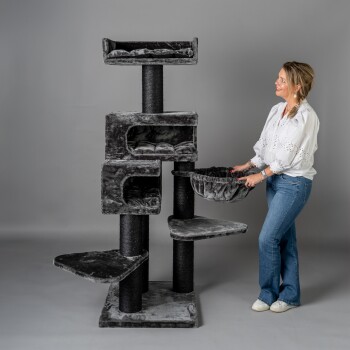 Eine Frau in einer weißen Bluse und blauen Jeans passt einen plüschigen grauen Katzenbaum mit mehreren Ebenen und gemütlichen Ruhebereichen an.