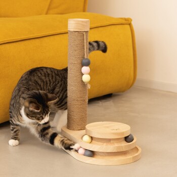 Eine verspielte getigerte Katze interagiert mit einem Holzspielzeug für Katzen, das einen Kratzbaum und bunte Bälle enthält, vor einem gelben Sofa.