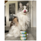 Eine flauschige Katze mit grau-weißen Fell leckt sich die Lippen, während sie neben drei Dosen Katzenfutter sitzt, die mit 'Lachs & Truthahn' beschriftet sind.