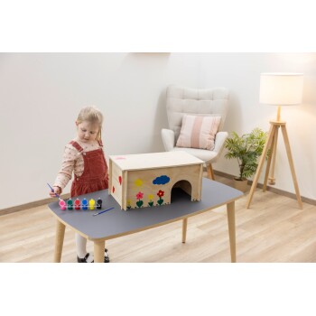 Kind malt ein hölzernes Haustierhaus mit bunten Blumen, Sonne und Wolke auf einem Tisch in einem gemütlichen Raum.