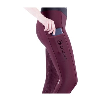 Nahaufnahme eines Frauenbeins in burgunderfarbenen Sportleggings mit "EQUIVA"-Logo, das ein Handy in die Seitentasche steckt.