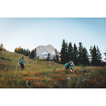 Eine Person in einer türkisfarbenen Jacke geht durch ein grasbewachsenes Feld mit einem Hund, der einen passenden türkisfarbenen Mantel trägt, vor einer Bergkulisse.