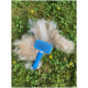 Eine blaue Haustierpflegebürste liegt auf einem Stück grünem Gras, umgeben von flauschigem, hellfarbenem Haustierfell und kleinen gelben Blumen.