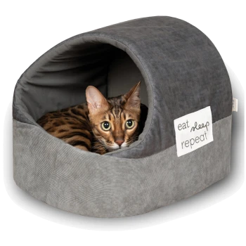 Ein gemütliches graues Katzenbett mit abgerundetem Oberteil und seitlichem Eingang, mit dem Text 'essen schlafen wiederholen' auf einem weißen Etikett.