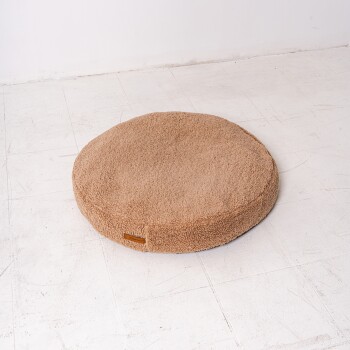 Rundes, plüschiges Haustierbett in hellbrauner Farbe, auf einem weißen Boden platziert, mit einem kleinen Etikett an der Seite.