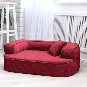 Rotes Haustierbett mit runder Form und zwei Seitenkissen, platziert in einem hellen Raum mit weißen Vorhängen und grünen Pflanzen.