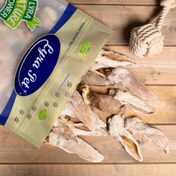 Eine Tüte Lyra Nature Power Haustierleckerlis mit getrockneten Kaninchenohren, die teilweise auf einer Holzoberfläche geöffnet ist, mit einem Seilspielzeug in der Nähe.