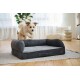 Fluffy tan-and-white dog mid-jump toward a dark gray bolstered dog bed on a rug in a bright living room