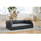 Flauschiger sandbraun-weißer Hund im Sprung auf ein dunkelgraues Bolster-Hundebett auf einem Teppich in einem hellen Wohnzimmer.