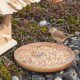 Ein kleiner brauner Vogel steht neben einem hölzernen Vogelhaus und pickt an einem Haufen Vogelfutter auf einem runden Holzbrett, umgeben von Kieselsteinen und Grünpflanzen.