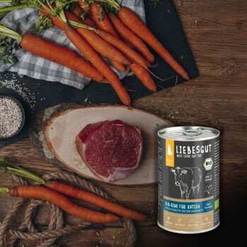 Dose mit der Aufschrift LIEBESGUT „BIO‑RIND FÜR KATZEN“ neben rohem Rindersteak und Karotten auf einem Holzbrett.