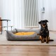 Ein kleiner schwarz-brauner Hund sitzt neben einem gemütlichen grauen Hundebett mit einem gelben Kissen in einem hellen, modernen Raum.