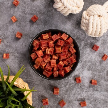 Schüssel mit kleinen, gewürfelten Haustierleckereien in einer schwarzen Schüssel, umgeben von verstreuten Leckereien, einem geflochtenen Korb mit grünem Gras und zwei Seilspielzeugen.
