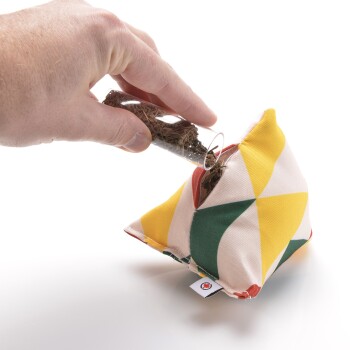 Eine Hand gießt lose Haustierbettung aus einem Glasrohr in einen bunten, geometrisch gemusterten Stoffbeutel, der für Haustiere entworfen wurde.