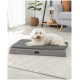 Ein kleiner weißer Hund ruht auf einem grauen Haustierbett mit plüschiger Oberseite, das auf einem weichen, hellen Teppich in einem modernen Wohnzimmer platziert ist.