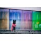 Ein schwarzer Hund, der einen leuchtend roten Regenmantel trägt, sitzt auf Steinstufen vor einem bunten, verschwommenen Glas-Hintergrund.